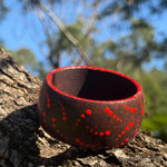 Tim Yule, Aboriginal Artist. A wide, chunky brown wooden cuff bracelet with red painted dots, representing Burning Starts, resting on a textured tree trunk outdoors. Side-Top View. Dot details around the rim