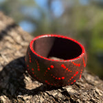 Tim Yule, Aboriginal Artist. A wide, chunky brown wooden cuff bracelet with red painted dots, representing Burning Starts, resting on a textured tree trunk outdoors. Top-View. Dot details around the rim
