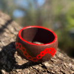 Tim-Yule-AboriginalAt-A dark brown wooden bangle with red circular pattern around the band, resting on a textured tree trunk. Sideview. Painted around the rim. Topview