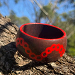 Tim-Yule-AboriginalAt-A dark brown wooden bangle with red circular pattern around the band, resting on a textured tree trunk. Sideview. Painted around the rim.