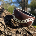 Tim-Yule-AboriginalAt-A dark brown wooden bangle with a white circular pattern around the band, resting on a textured tree trunk.- CloseupView