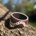 Tim-Yule-Aboriginal Art- A dark brown wooden bangle with a white circular pattern around the band, resting on a textured tree trunk. TopSide view.