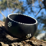 Tim Yule, Aboriginal Artist. A wide, chunky black wooden cuff bracelet with white dotted trim and white dotted floral pattern, resting on a textured tree trunk outdoors ranches in the background. Side view.