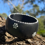 Tim Yule, Aboriginal Artist. A wide, chunky black wooden cuff bracelet with white dotted trim and white dotted floral pattern, resting on a textured tree trunk outdoors ranches in the background. Side