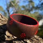 Tim Yule, Aboriginal Artist. A wide, chunky brown wooden cuff bracelet with white dotted trim and red dotted floral pattern, resting on a textured tree trunk outdoors ranches in the background. Side top view.
