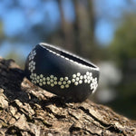 A wide black  wooden bangle with a white dotted flowers in pattern, resting on a textured tree trunk with. Close up