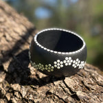 A wide black  wooden bangle with a white dotted flowers in pattern, resting on a textured tree trunk with. Topview
