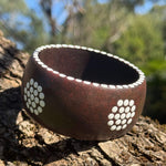 Tim Yule, Aboriginal Artist. A wide, chunky dark brown wooden cuff bracelet with white circular dot patterns representing meeting places on the sides, resting on a textured tree trunk. dot details around the rim. Top