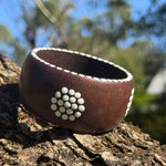 Tim Yule, Aboriginal Artist. A wide, chunky dark brown wooden cuff bracelet with white circular dot patterns representing meeting places, resting on a textured tree trunk. dot details around the rim. SideClose up