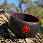 Tim Yule, Aboriginal Artist. A wide, chunky black wooden cuff bracelet with red circular dot patterns representing meeting places on a burned land. pattern on the sides, resting on a textured tree trunk. dot details around the rim. Close up