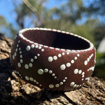 Tim Yule, Aboriginal Artist. A wide, chunky brown wooden cuff bracelet with white painted dots, representing Showers of stars, resting on a textured tree trunk outdoors. Side-View. Dot details around the rim