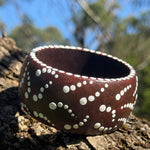 Tim Yule, Aboriginal Artist. A wide, chunky brown wooden cuff bracelet with white painted dots, representing Showers of stars, resting on a textured tree trunk outdoors. Side-Closeup-View. Dot details around the rim